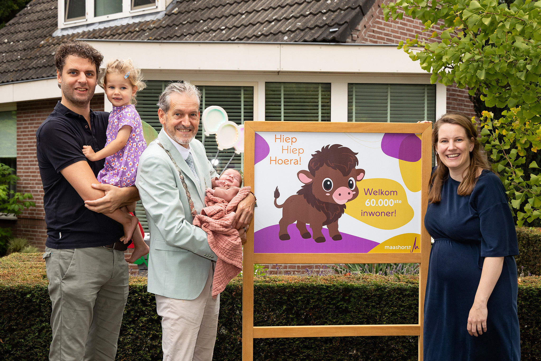 Burgemeester houdt een pasgeboren baby in zijn armen. Naast hem staan de trotse ouders met hun peuter. Ze poseren samen bij een bord met een vrolijke illustratie van een wisent en de tekst: "Hiep Hiep Hoera! Welkom 60.000ste inwoner!"